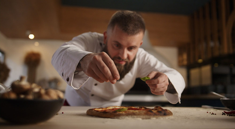 Image of chef sprinkling fresh basil on a cooked pizza in a restaurant.