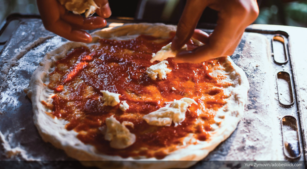 Image of pizzeria employee adding cheese to a pizza.