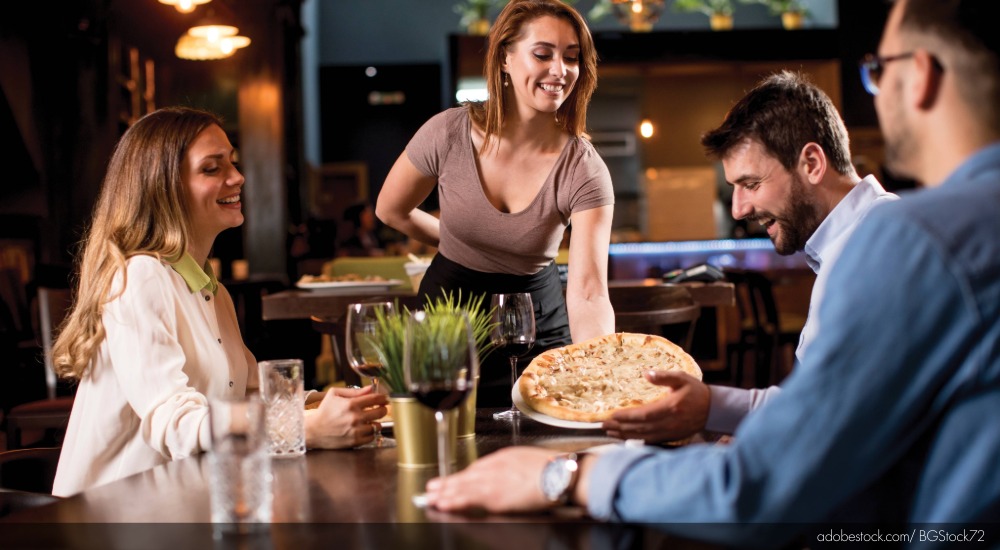 Image of a server delivering a pizza to a table at a restaurant.